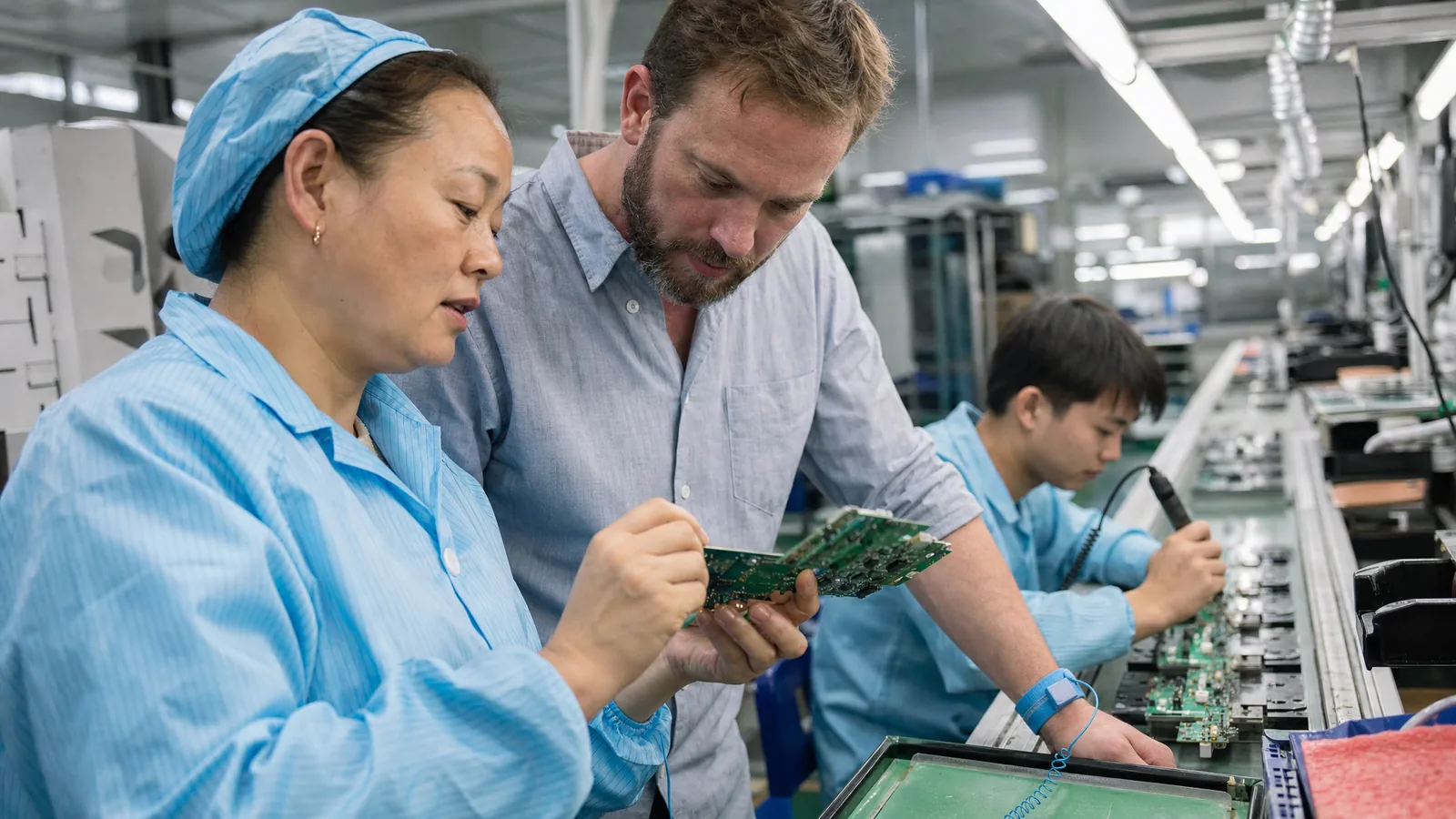A production manager on a Shenzhen electronics line shows a circuit board to an American buyer, who leans in to look closely